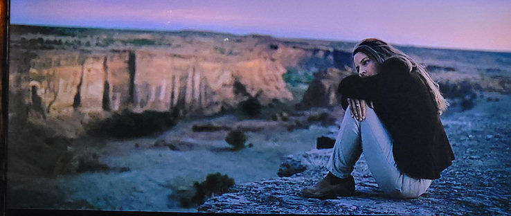 Person lost in thought near the edge of a cliff, watching the sun set over a red sandstone canyon. A greenish tinted river is in the distance.