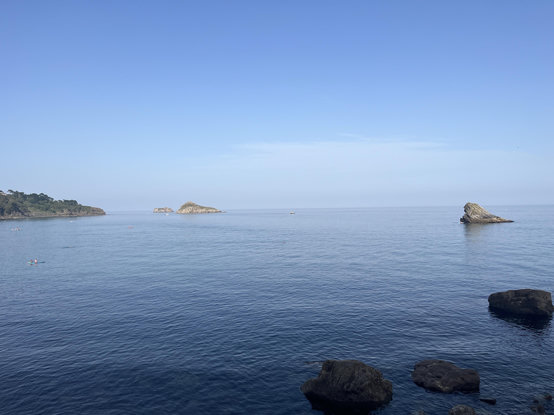 View from Meadfoot Beach with the horizon just under half way up the picture, the sea and sky are both light blue with the hint of low sea cloud in the far distance. On the left hand horizon is Thatcher Point, then just off-shore in the distance is Orestone Rock with Thatcher Rock being nearer. Finally, Shag Rock is on the right hand horizon. The sea is very calm and there are some small rocks protruding in the bottom right of the picture.