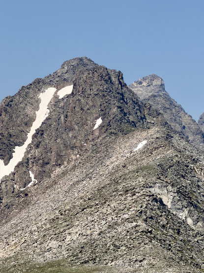 A rocky mountain landscape featuring steep cliffs, boulders, and patches of snow under a clear blue sky.