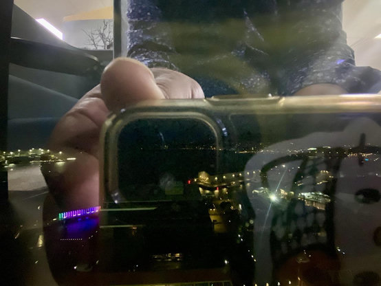 A reflection of a hand holding a phone up to an office window to take a picture across Wellington Harbour at dawn. The sun is obscured by low cloud. Lights are visible around the waterfront and city.