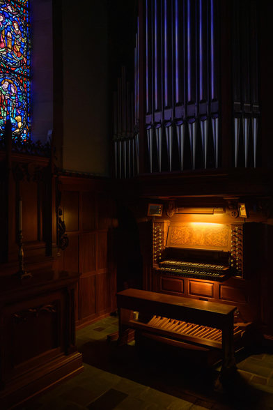 A photo of an organ console with the lights on in an otherwise dark space. A stained glass window at upper left is reflecting in the organ pipes.