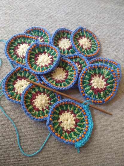 Twelve circular crochet motifs with a white and burgundy centre, and a ring of two tone green, then blue with orange bead detail.