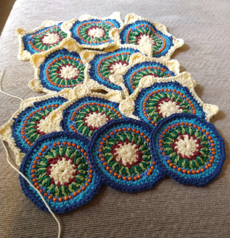 Twelve circular crochet motifs with a white and burgundy centre, and a ring of two tone green, then blue with orange bead detail, the top nine have white star like edging