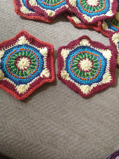 Two hexagonal crochet motifs with a white and burgundy centre, and a ring of two tone green, then blue with orange bead detail, on the left has an orange border