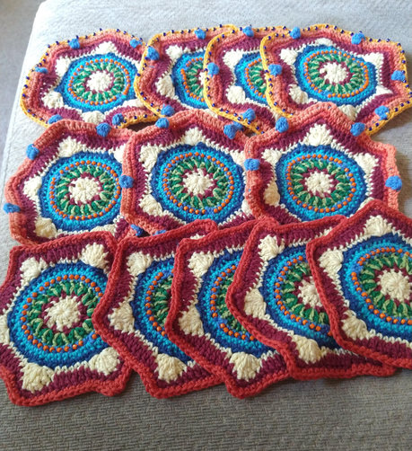 Twelve hexagonal crochet motifs with a white and burgundy centre, and a ring of two tone green, then blue with orange bead detail.
At the top are five hexagon, with bobble and bead detail on the edge round