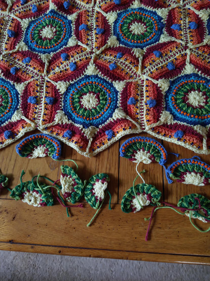 Part section of hexagonal crochet project with a white and burgundy centre, and a ring of two tone green, then blue with orange bead detail
At the bottom are partly finished half hexagons