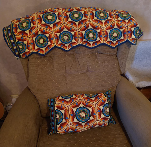 Multi coloured Crochet piece laid across the back of a chair on the seat if the chair is a cushion in the same pattern