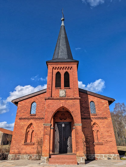 A small neo-Gothic red brick former church in Schmerwitz, built in 1898, now serving as an event and wedding venue for Schmerwitz Estate. The building features a tall, slender pointed dark slate spire with a decorative finial, three pointed arched windows with tracery, and a central pointed arched entrance with double dark wooden doors. The red brick facade includes stone detailing around the windows and a stone base course, with a short flight of steps leading to the entrance. Trees are visible in the background, and the structure stands against a bright blue sky with scattered white clouds.