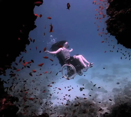 Photo featuring a woman with arms out in a specially powered wheelchair with oxygen equipment, under the sea among fishes in blue water