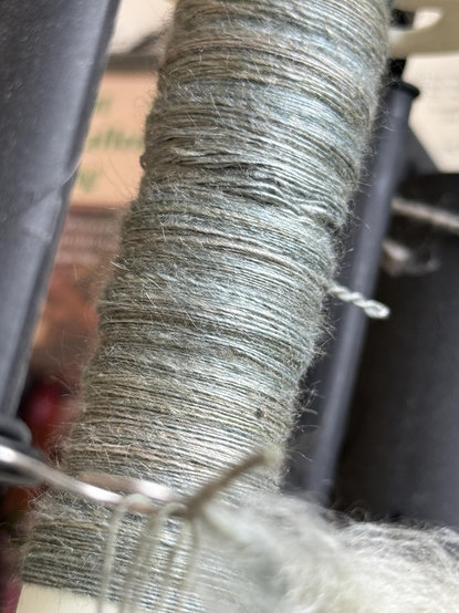 A silvery green-blue yarn singles wrapped around an e-wheel bobbin. There’s a pigtail of singles sticking out of one side from when I was having issues