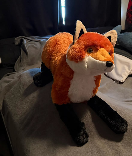 Big red fox plush, 30" in size, similar in size to the larger blahaj. It is laying on a large blahaj shaped lump on our bed.