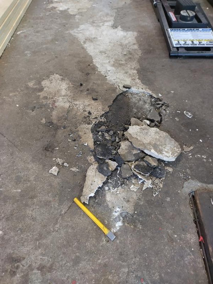 Valokuva: vanha läikikäs ja moneen kertaan paikkailtu betonilattia, osa paikkauksesta piikattuna auki, paikoin isoina laattoina.