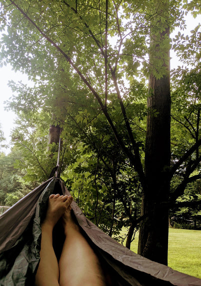 Looking down towards my legs and feet as I am nestled and slightly swinging in a forest green and grey hammock among some deciduous trees in my yard. Hazy cloudy skies.