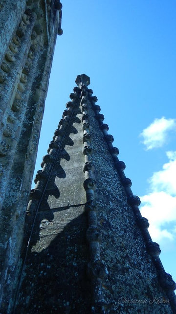 Close-up view of a weathered stone church spire with decorative protrusions, seen from below against a blue sky with a few clouds.