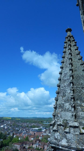 Stone church spire with decorative details in the foreground, overlooking a town with houses and buildings under a blue sky with scattered clouds.