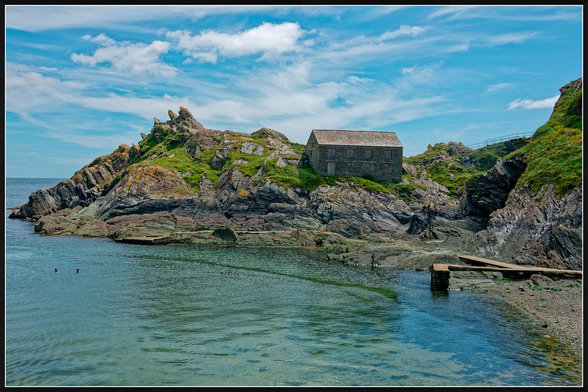 "A quiet Cornish cove at Polperro featuring turquoise water lapping against rocky shores lined with brown seaweed. A solitary stone cottage sits atop a grassy rock formation jutting into the bay, with wooden planks forming a small jetty extending into the calm water under a bright blue sky dotted with fluffy white clouds."

