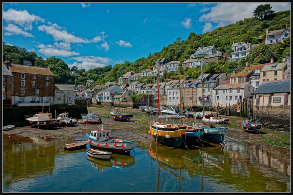 "Traditional Cornish fishing harbour at Polperro with colourful fishing boats resting in shallow tidal waters. Wooden hulls reflect in the calm green-blue water while tightly packed cottages and houses climb steeply up the wooded hillside behind. A bright blue sky with scattered white clouds completes the picturesque scene."