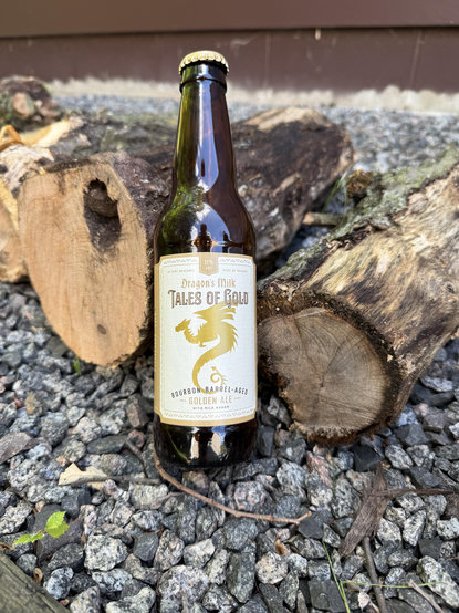 A brown bottle with a white label has gold lettering and a dragon in silhouette. it is leaned against some cut oak and birch wood on a bed of crushed granite stones