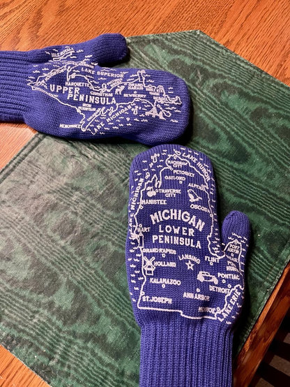 A pair of blue mittens featuring a map design of Michigan's Upper and Lower Peninsulas. Michigan’s upper and lower peninsulas are shaped like mittens to begin with. The mittens display various cities and landmarks in white rubber lettering, resting on a table.
