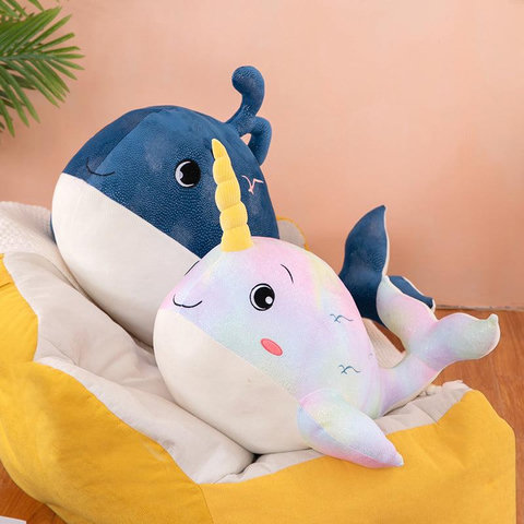 a dark blue whale plushie sits behind a pastel, rainbow-colored narwhal plushie with a yellow horn, both resting on a yellow and white cushioned surface.