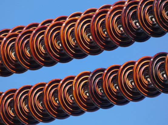 Photo of a vivid blue sky across which stretches a pair of power lines completely covered in bloodred ceramic rings like flat, glossy, ringed beads, the coils of an 1980s telephone cord, or a laundry line of fiestaware hung out to dry.