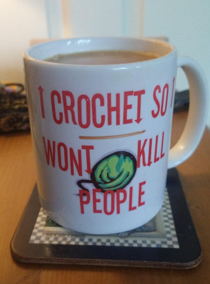 Mug of tea on a bedside cabinet with red text saying I crochet so I won't kill people it has a horizontal crochet hook in the middle above a ball of green yarn