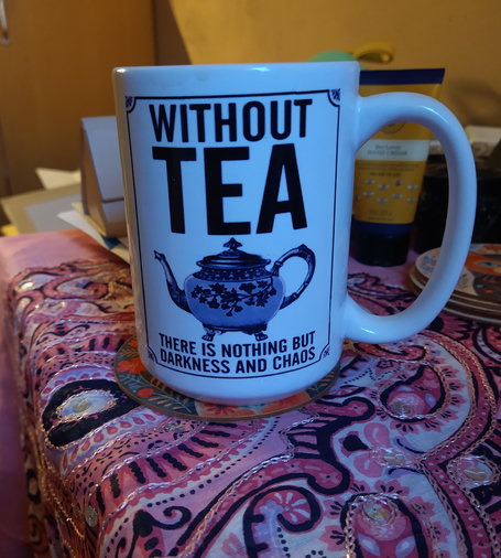 Mug of tea on a small coffee table covered with a pink cloth, on the mug are the words without Tea there is nothing but darkness and chaos, there is a pink tea pot in the middle of the mug