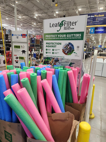 Photo taken in a big box hardware store. A sign advertises gutter leaf filters. Immediately in front our boxes of brightly colored foam pool noodles.