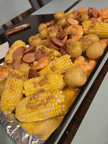 A cookie sheet covered in foil. On it is corn on the cob, sausages, baby potatoes, and shrimp all covered in cajun seasonings, old bay, and garlic butter