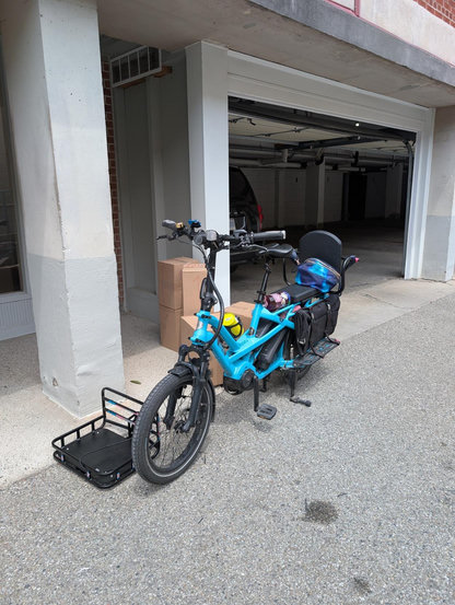 Blue long-tail cargo bike in front of a building garage, with a front rack on the ground and a stack of boxes behind