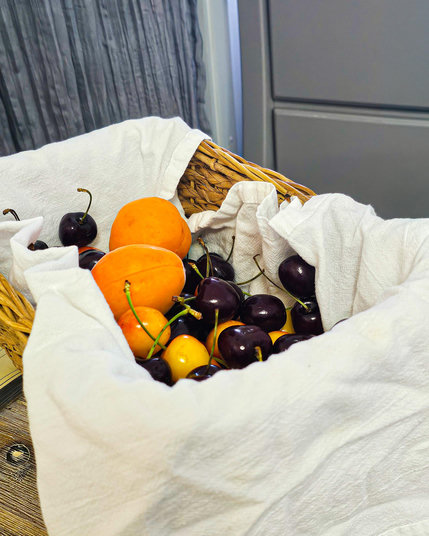 A  lined basket with a few pounds of Rainier and Bing cherries and a couple of lush apricots sits on a coffee table. There is a gray chest of drawers and a gray drape in the background. Rainier cherries are yellow with a red blush and are soft and sweet. Bing cherries are dark dark red, firm and full of good cherry flavor. The apricots are very large! But can't hold a candle to the small ones I used to grow in Colorado