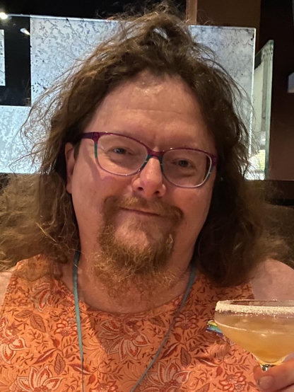 miriam, a year before egg crack, wearing an orange top and a goatee, with frankly embarrassingly messy hair. her eyes are squinty and her smile hollow. she's tired and scruffy.