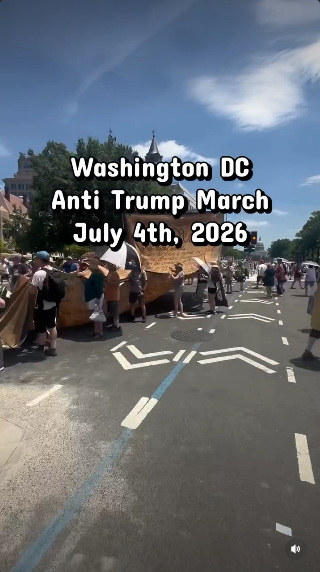 A big crowd of people is carrying a long tan-colored copy of the Constitution through the streets of DC under a blue sky. This is the end of the march where the "We the People" are visible at the top.