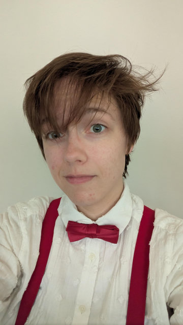Daneel, a white genderqueer person with floppy brown hair and green eyes, dressed in a white dress shirt and red bowtie and braces. Their hair is messy, sticking up in some places and in their eyes in others. They're smiling a little at the camera.