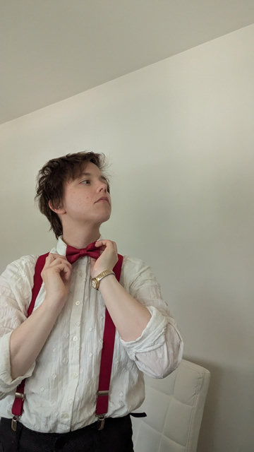 Daneel, standing away from the camera. They're looking off to the side at an unseen mirror, adjusting their bowtie.