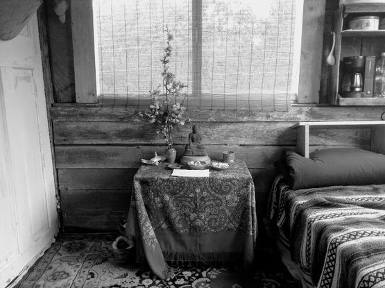 An old TV table covered with a shawl makes an altar (inside it is pantry). On the eight is cot, blanket, seiza bench, and on the wall the little crate kitchen. On the altar a tall branch o flowering quince complements the volcanic-ash Buddha.