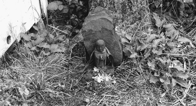 Jizo (Kshitigarbha) backed by a stone from the dry wash, back by the stump of the former shade tree, with vinca blossoms at his feet. Wall of hut visible at left.