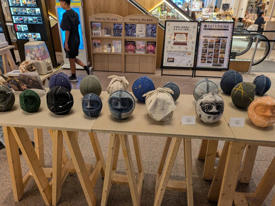 A line of unpainted daruma dolls on a makeshift wooden table in a mall area