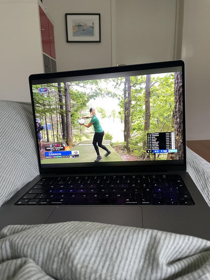 En macbook ovanpå täcket på min mage i sängen. På skärmen syns en discgolfspelare kasta ut en frisbee från en tee.