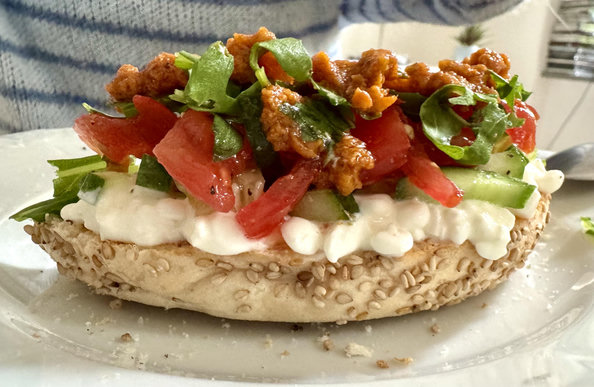 Auf einem weißen Teller liegt ein aufgeschnittener Bagel mit einem Belag aus weißem Hüttenkäse und weiterem roten und grünen Belag aus Tomaten, Gurken, Pesto und so weiter.