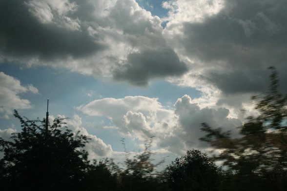 Foto im Querformat. Blick aus einem fahrenden Zug. Unten sind grüne Büsche, darüber ein Stück blauer Himmel, größerenteils verdeckt durch weiß-graue Wolken. Aufgrund der Fahrgeschwindigkeit ist das Bild leicht unscharf.