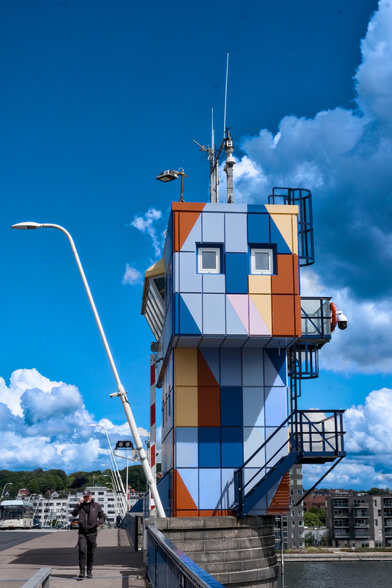 Neben einer Brücke steht ein farbenfroher, moderner Aussichtsturm mit geometrischen Mustern. Eine schwarz gekleidete Person geht auf dem Weg in der Nähe des Turms. Der Himmel ist teilweise bewölkt, mit leuchtenden Blautönen.