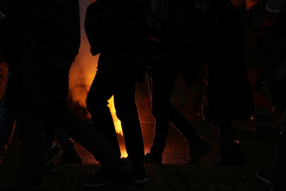 Foto im Querformat. Es ist dunkel wie bei einer Nachtaufnahme. Vor dem gelb leuchtenden Bengalo sind die Silhouetten von Menschen zu erkennen. Der Hintergrund ist orange. Fotografiert wurde leicht nach unten.