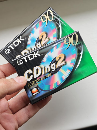 Ein Foto von zwei originalverpackten alten Audio-Kassetten "TDK CDing2", die schwarz-grün akzentuierte Folien-Verpackung zeigt eine schillernde CD. Gehalten in meiner Hand, weißer Hintergrund.