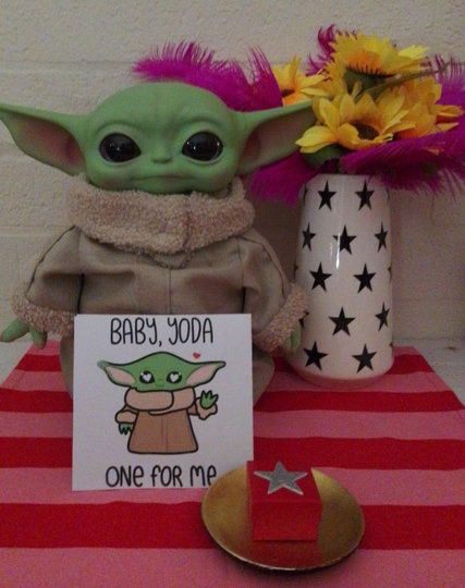 Baby Yoda doll, looking at the camera, while standing next to some some sunflowers and pink feathers in a white vase with black stars on it. Propped up against Baby Yoda is a white card with a pic of Baby Yoda with heart eyes, and the black text that says "Baby, Yoda One For Me." Next to the card is a round gold plate with a red ring box with silver star on top. Everything sits on a bright red and pink striped mat.