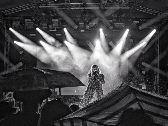 Black-and-white photograph of Mieze Katz, lead singer of German band MIA, performing at the centre of a rain-soaked stage. Dramatic beams of stage lighting cut through the darkness, illuminating her glittering costume as water droplets spray around her. In the foreground, silhouetted audience members hold umbrellas aloft. The stage features a visible metal truss structure with professional lighting fixtures, creating a striking contrast between the brightly lit performer and the shadowy, umbrella-filled crowd.